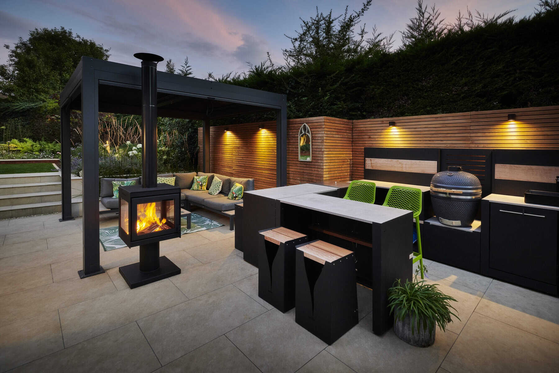 Covered outdoor seating area with a wood-burning stove and modern outdoor kitchen on a paved patio at dusk.